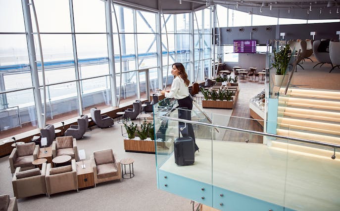 Woman standing in modern airport lounge with suitcase, overlooking seating area.