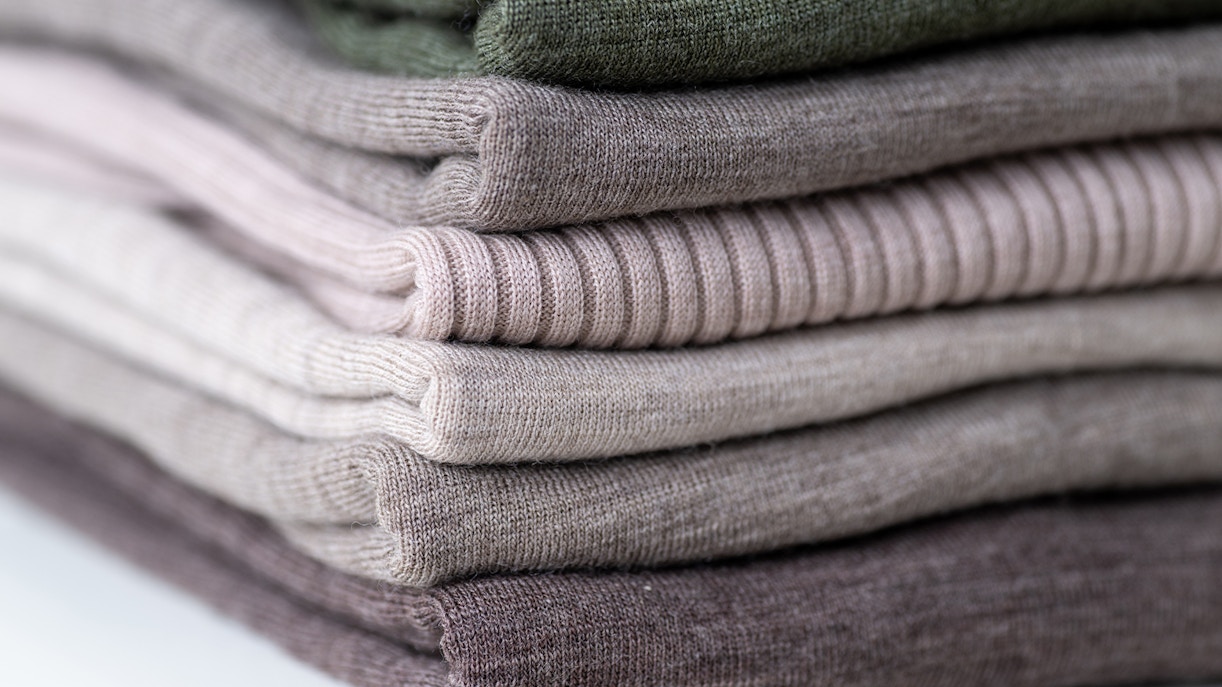 Stack of folded Merino wool sweaters in various neutral colors.