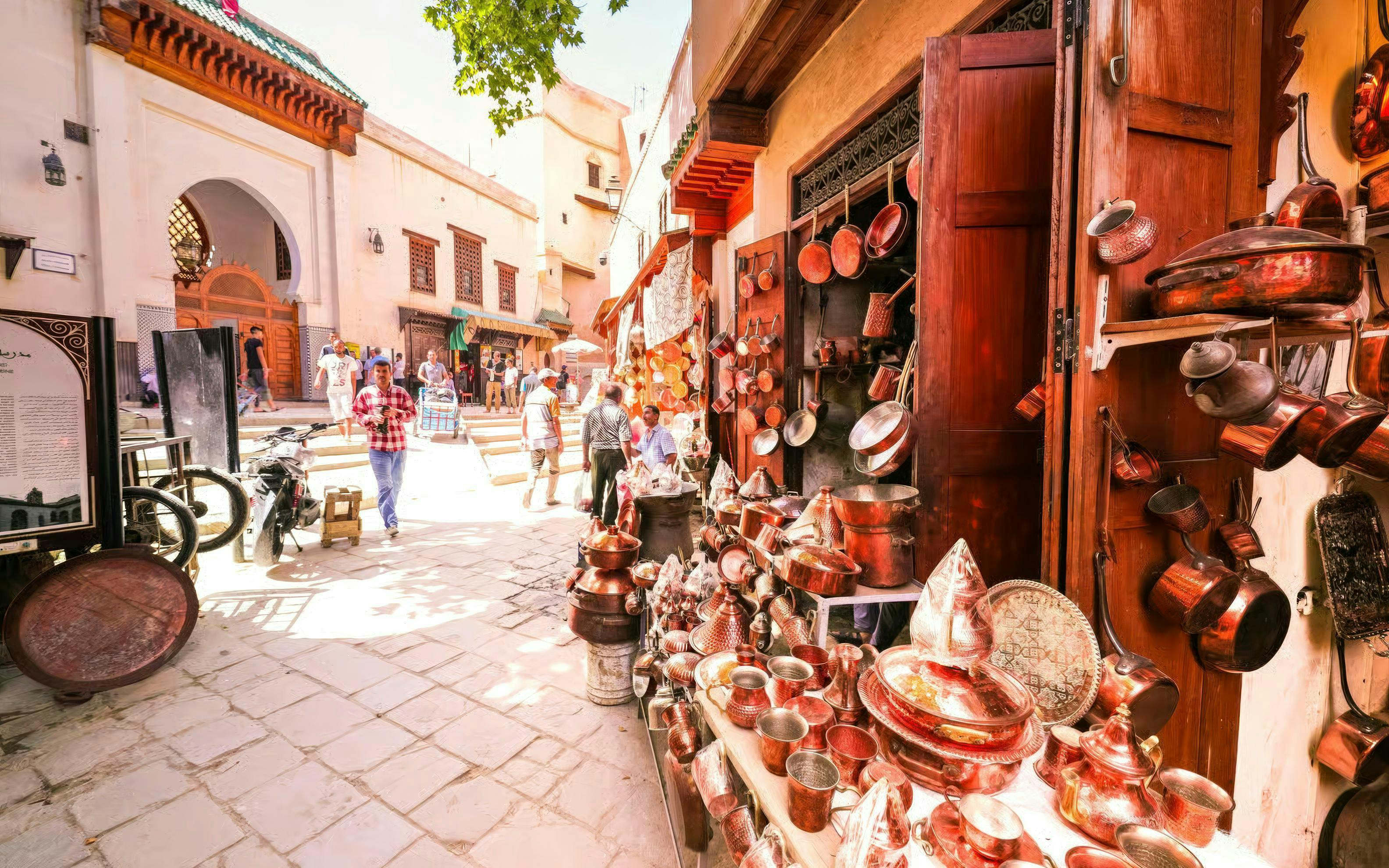 Copperware shop in Fes Medina with people walking near Al Attarine Madrasa entrance.