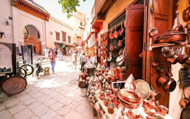 Copperware shop in Fes Medina with people walking near Al Attarine Madrasa entrance.