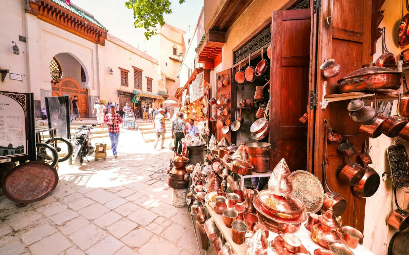 Copperware shop in Fes Medina with people walking near Al Attarine Madrasa entrance.