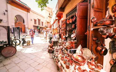 Copperware shop in Fes Medina with people walking near Al Attarine Madrasa entrance.