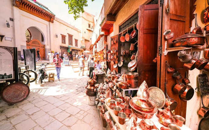 Copperware shop in Fes Medina with people walking near Al Attarine Madrasa entrance.