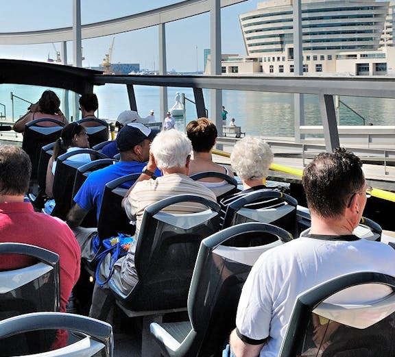 Tourists on a hop-on hop-off bus in Barcelona with waterfront view.
