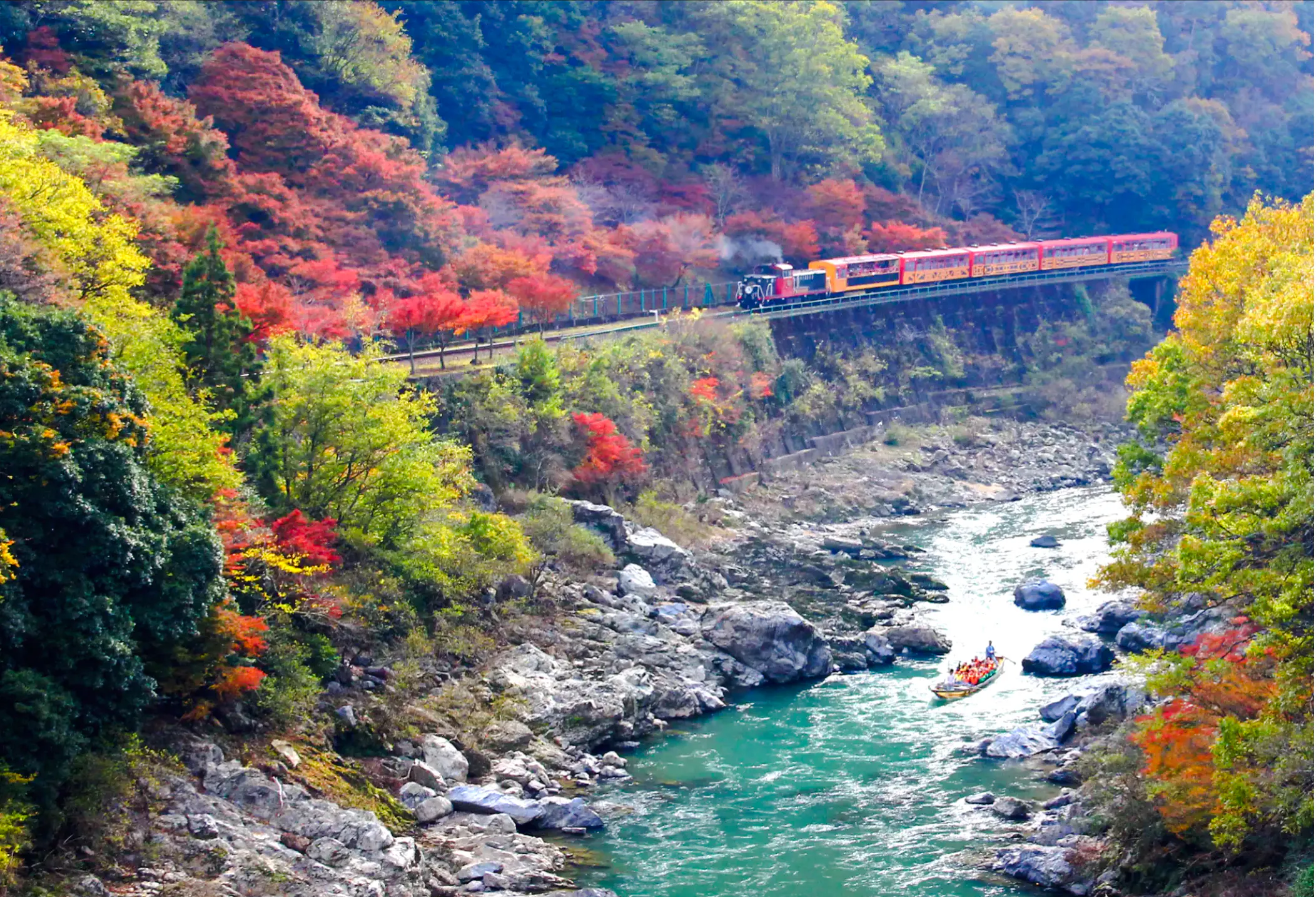 Train passing through colorful autumn forest along Hozugawa River, Kyoto, with a boat on the water.