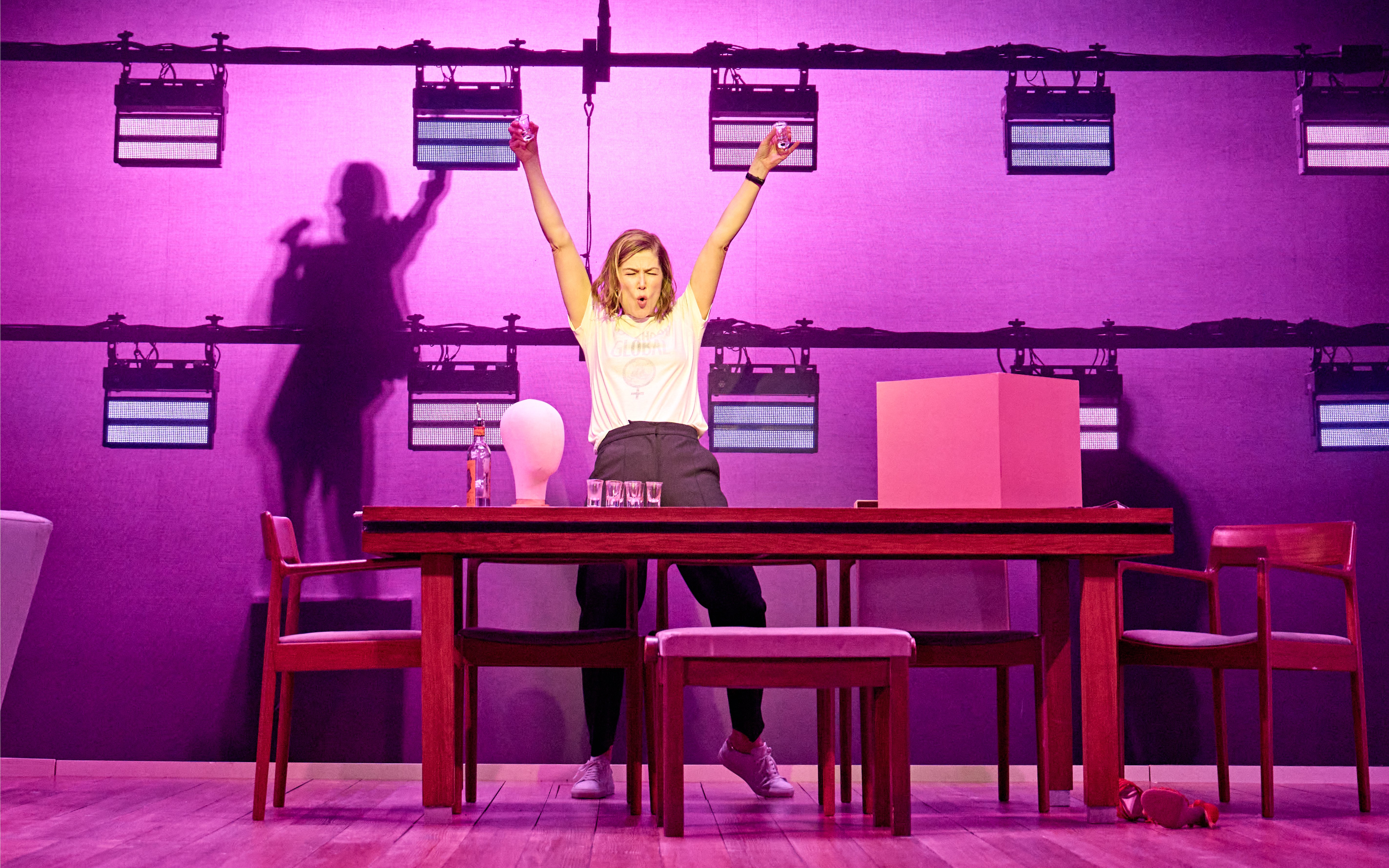 Woman celebrating with raised arms behind a table on stage in a theatrical performance.