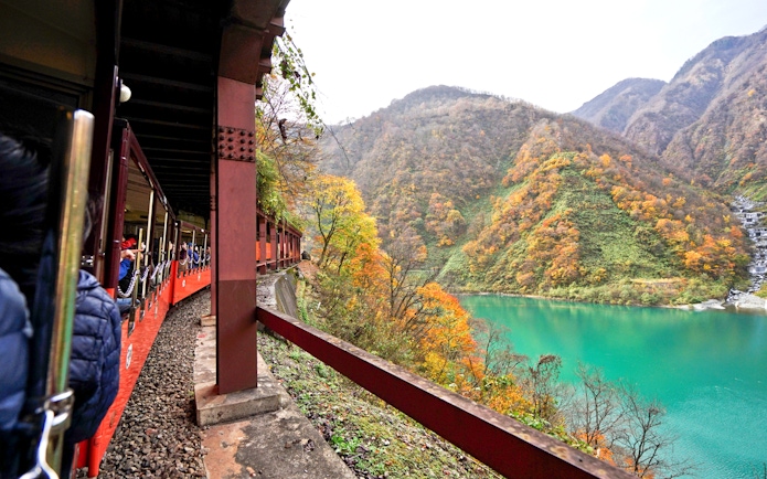 Train journey through Hokuriku mountains with autumn foliage and turquoise river.