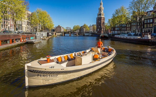 Amsterdam canal cruise boat with drinks, passing historic buildings and Montelbaanstoren tower.