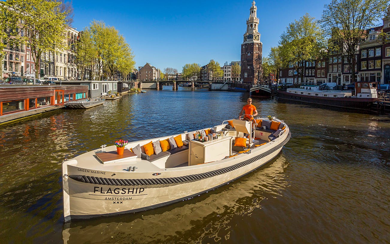 Amsterdam canal cruise boat with drinks, passing historic buildings and Montelbaanstoren tower.