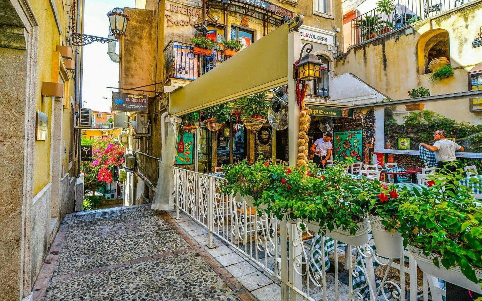 Taormina street with outdoor café, colorful flowers, and historic buildings.