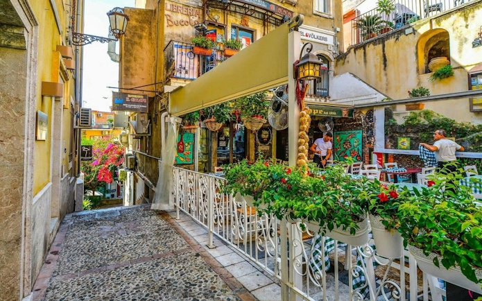 Taormina street with outdoor café, colorful flowers, and historic buildings.