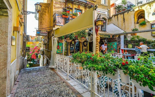 Taormina street with outdoor café, colorful flowers, and historic buildings.