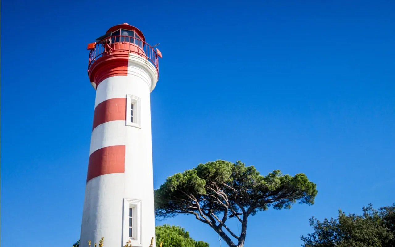 Lighthouse with red and white stripes in La Rochelle, France, near a tree under a clear blue sky.