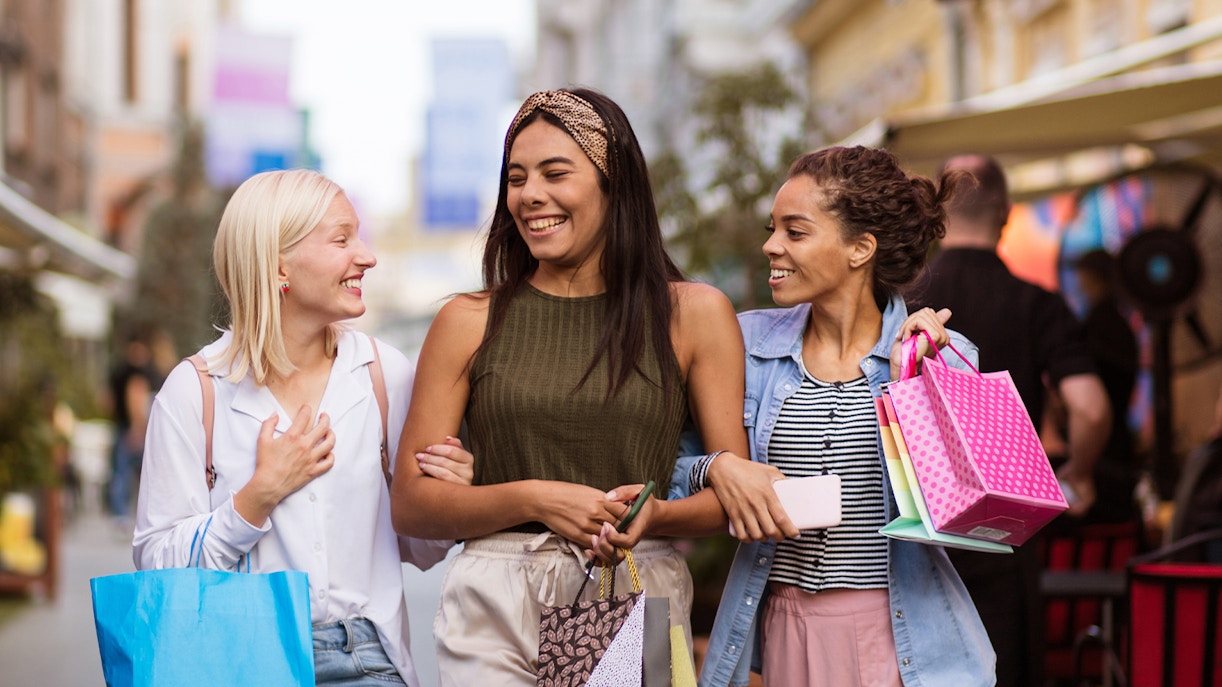 Women shopping on a lively street, carrying colorful bags and smiling.