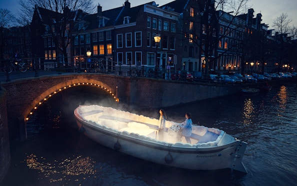 Cloud Boat Cruise on Amsterdam canal at dusk with illuminated bridge.