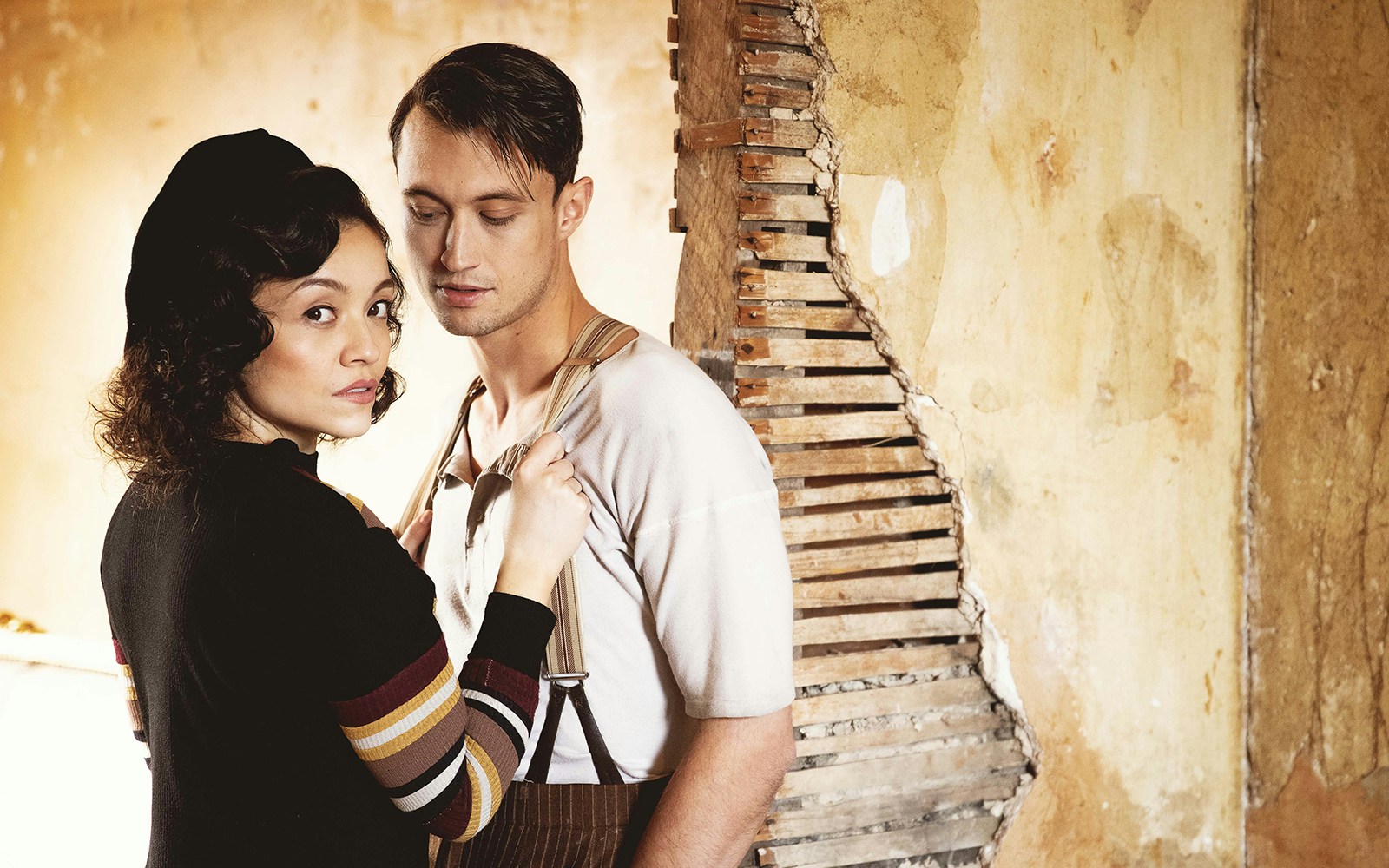 Couple in vintage attire standing by a damaged wall, evoking Bonnie & Clyde theme.