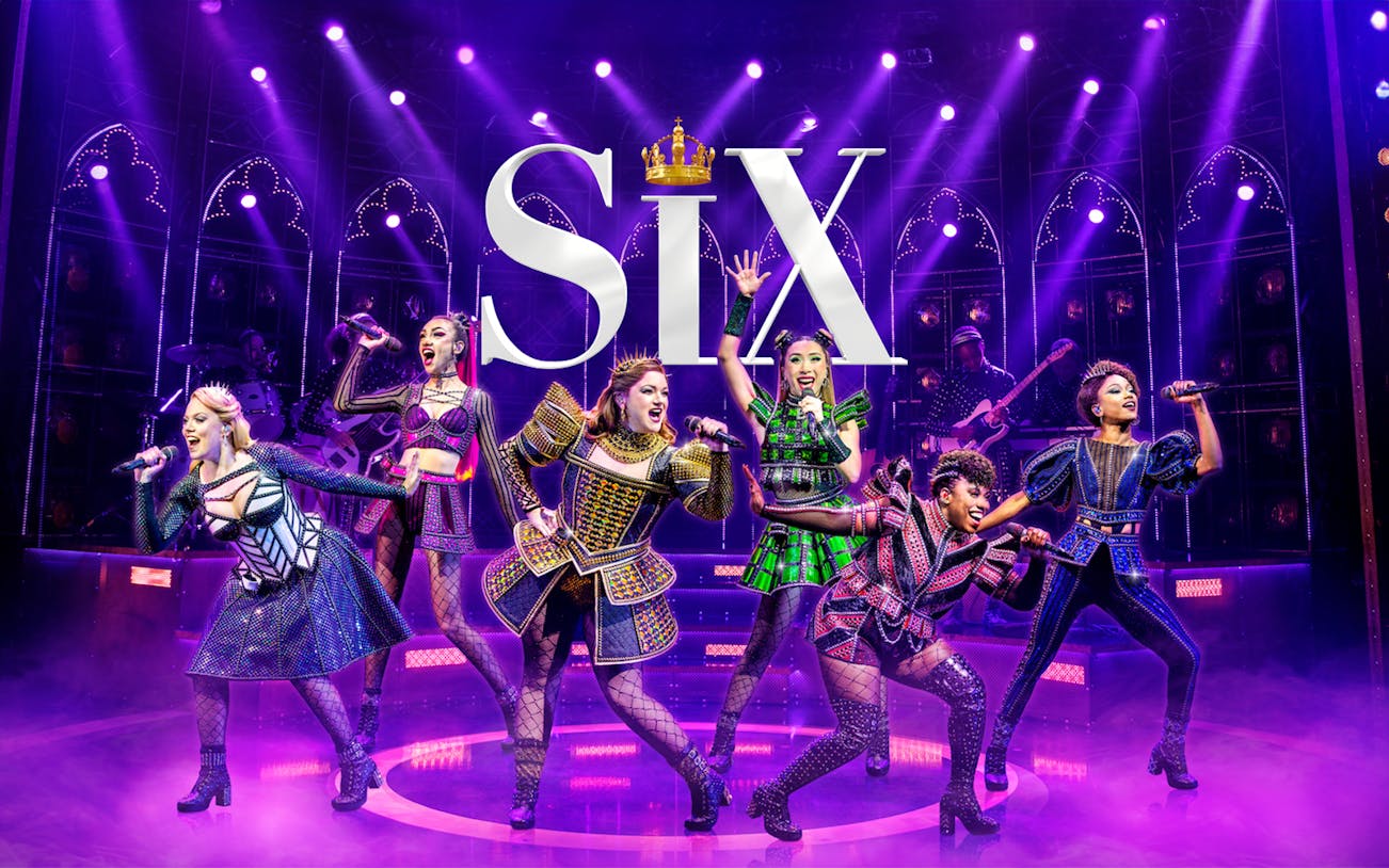 Performers on stage in vibrant costumes during the musical "Six" with dramatic lighting.