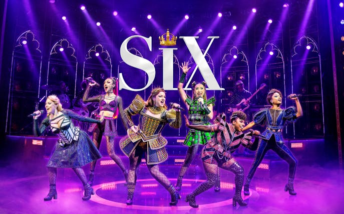 Performers on stage in vibrant costumes during the musical "Six" with dramatic lighting.