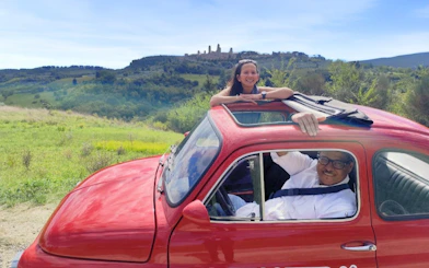 Vintage Fiat 500 driving through Chianti countryside with vineyards in the background.