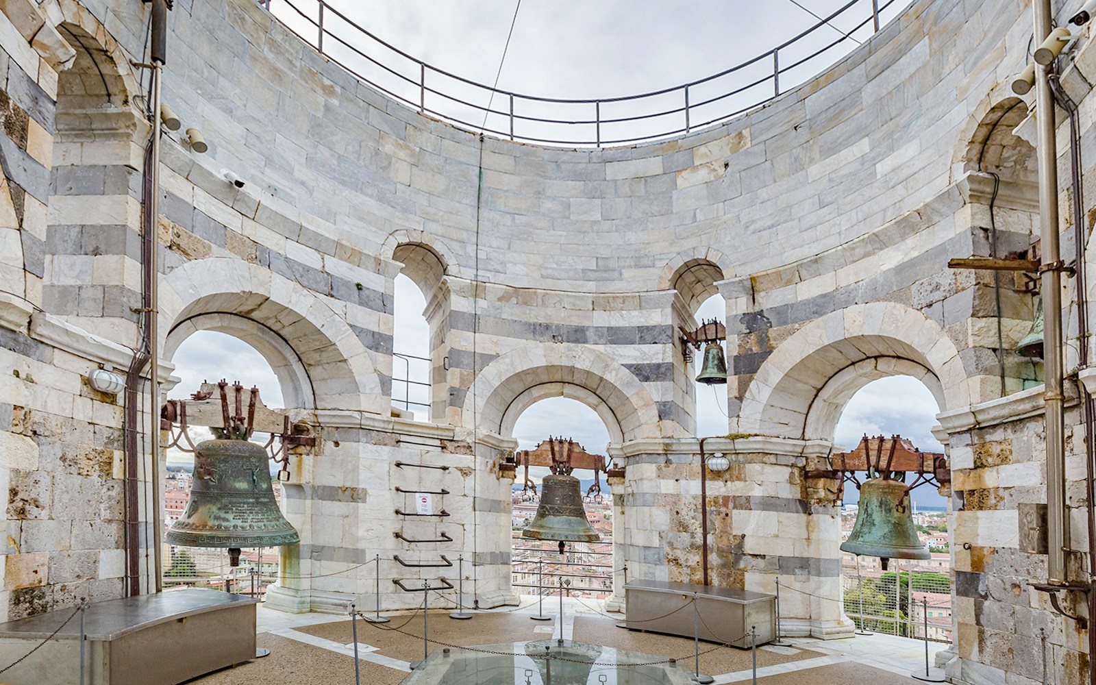 Bells at the top of the Leaning Tower of Pisa, Italy, with city view through arches.