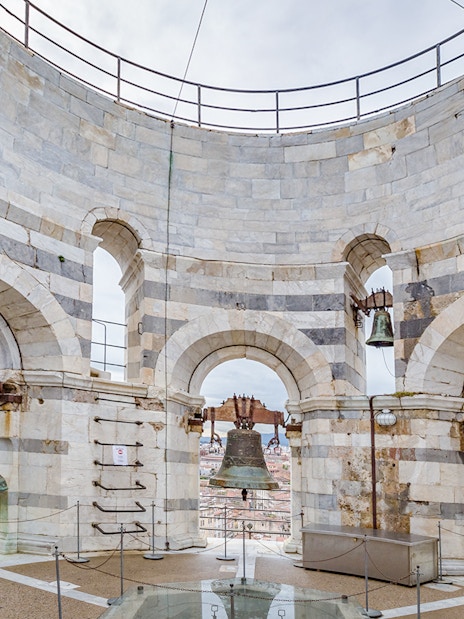 Bells at the top of the Leaning Tower of Pisa, Italy, with city view through arches.