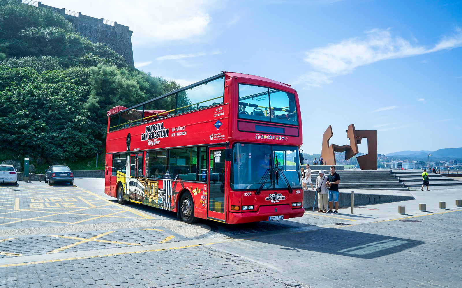 Red double-decker tour bus in San Sebastian near a modern sculpture and scenic view.