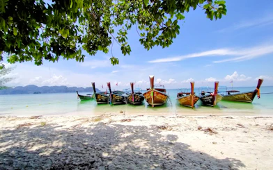 Longtail boats anchored on a sandy beach in Krabi, Thailand, with distant islands visible.