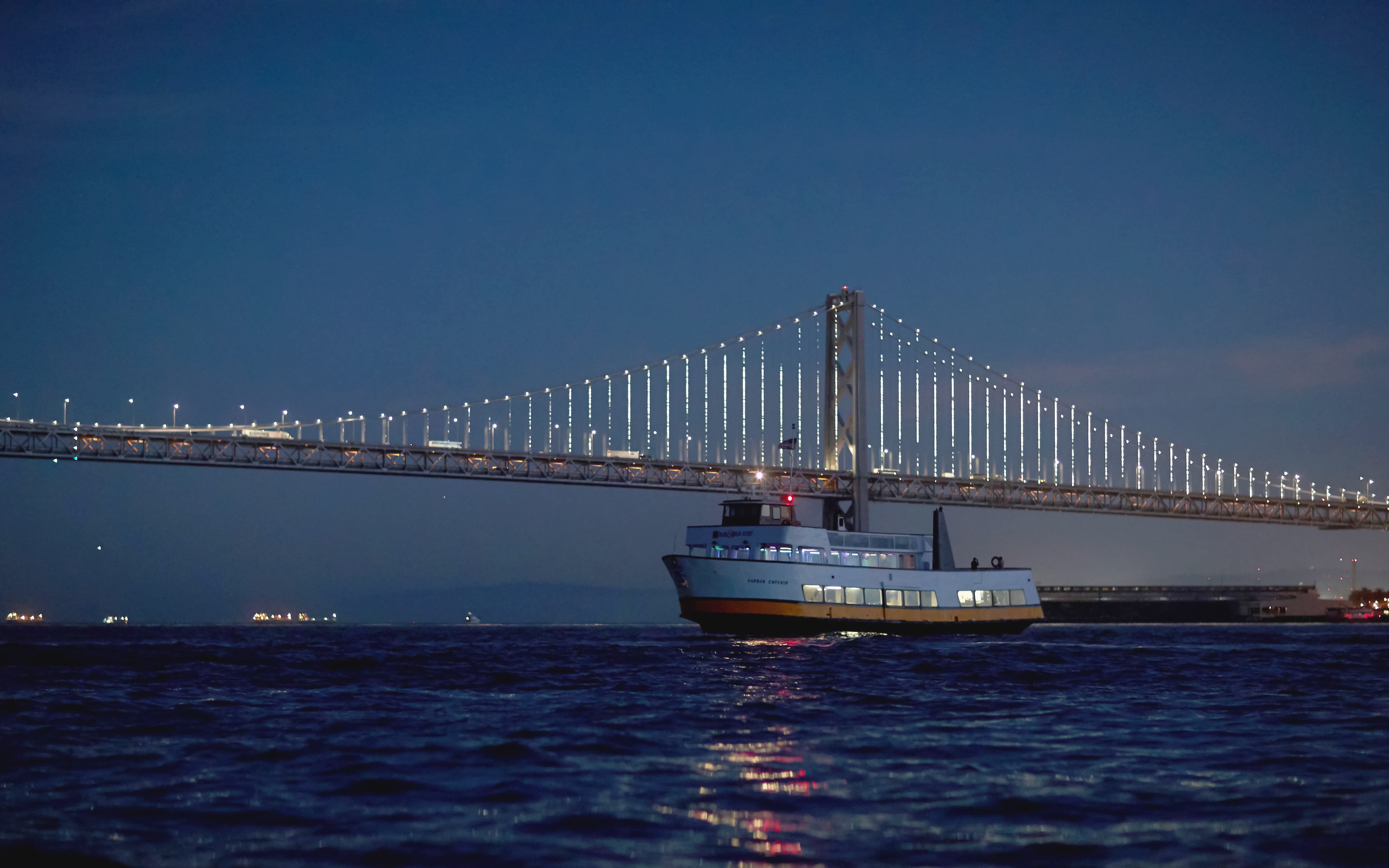 Cruise boat sailing under illuminated Bay Bridge at dusk, San Francisco Bay.