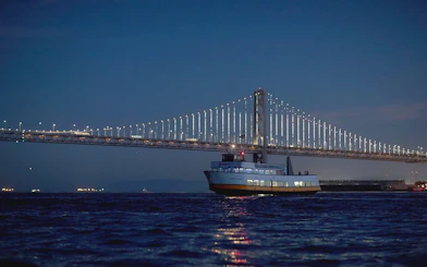 Cruise boat sailing under illuminated Bay Bridge at dusk, San Francisco Bay.