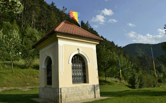 Small historical building with Romanian flag in Schei, Brașov, surrounded by lush greenery.