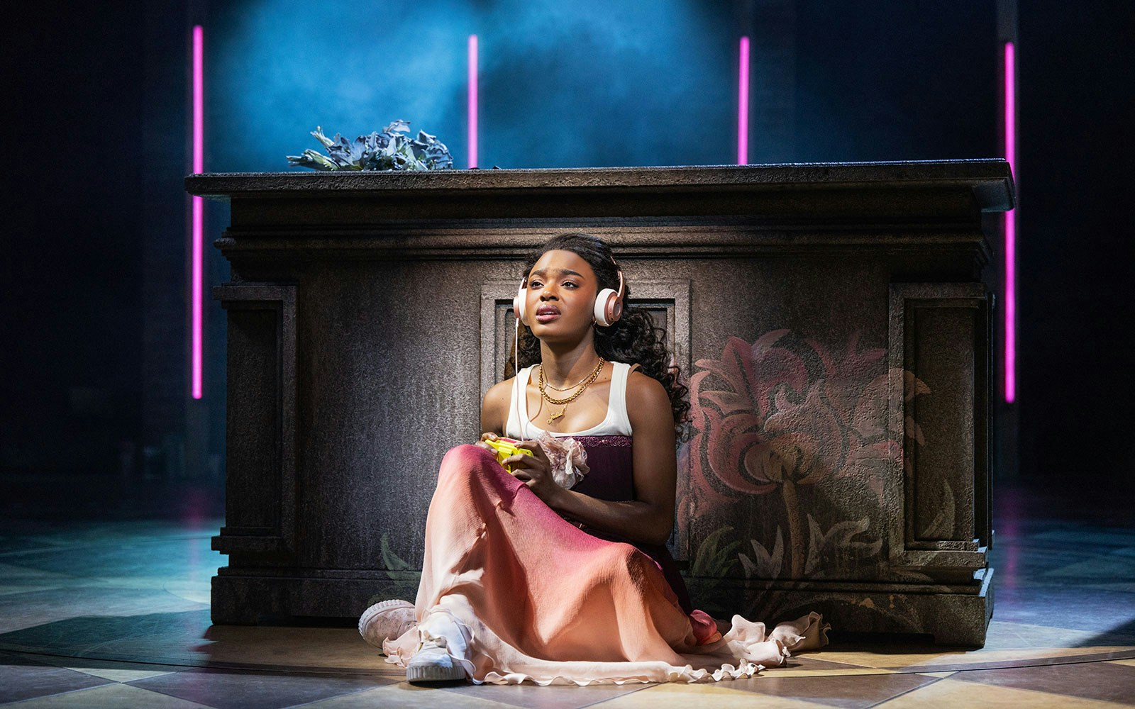 Performer in a scene from "& Juliet" musical, sitting by a decorated stone structure.
