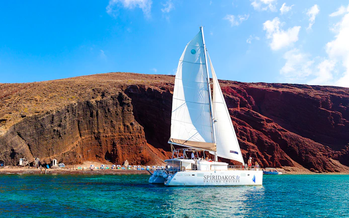 Catamaran sailing near the red cliffs of Santorini, Greece.