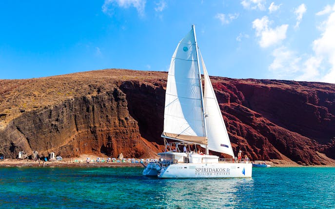 Catamaran sailing near the red cliffs of Santorini, Greece.