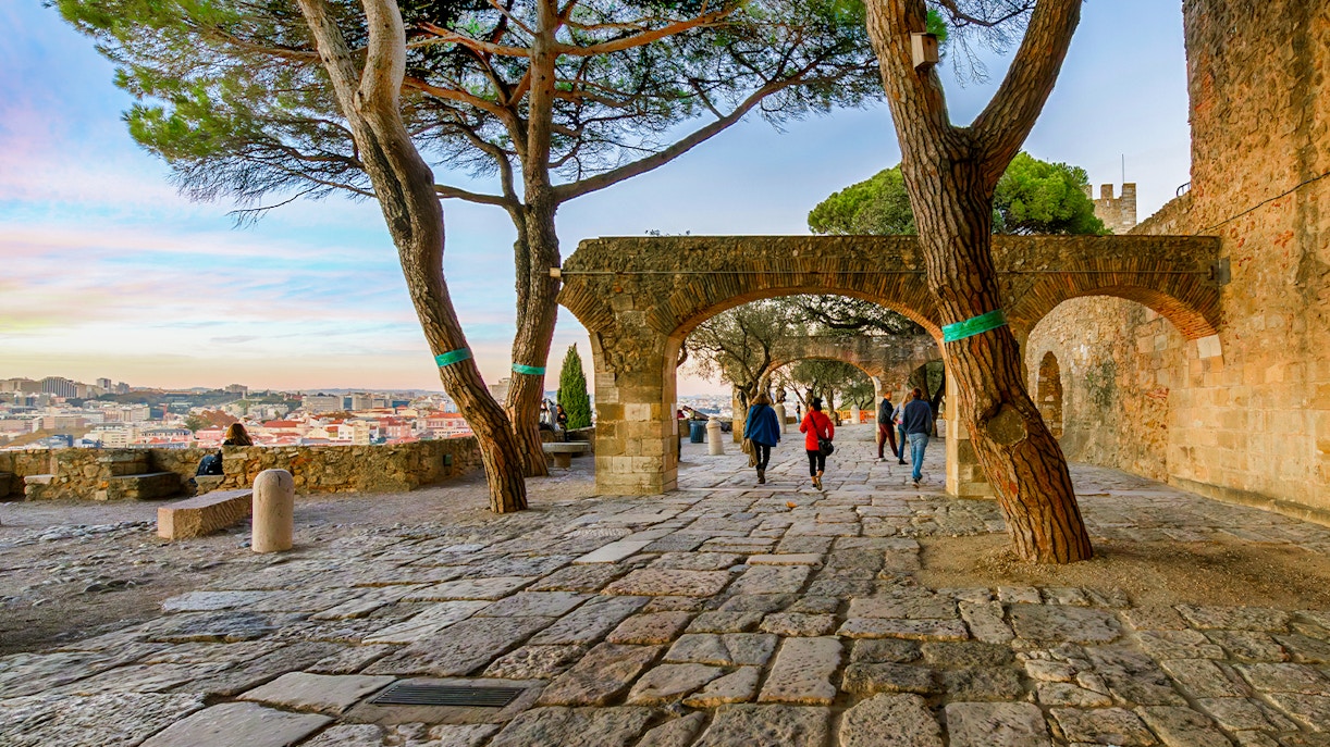Visitors walking on the terrace of Saint Georges Castle, Lisbon, with views of Alfama and Baixa at sunset.