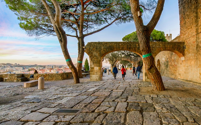 Visitors walking on the terrace of Saint Georges Castle, Lisbon, with views of Alfama and Baixa at sunset.