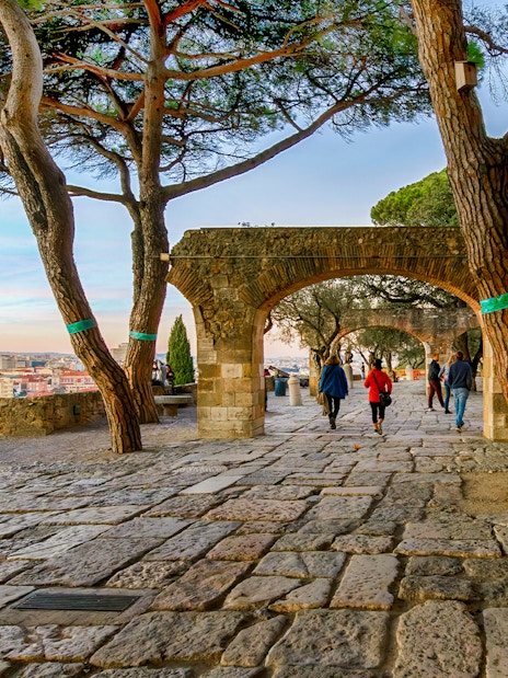 Visitors walking on the terrace of Saint Georges Castle, Lisbon, with views of Alfama and Baixa at sunset.