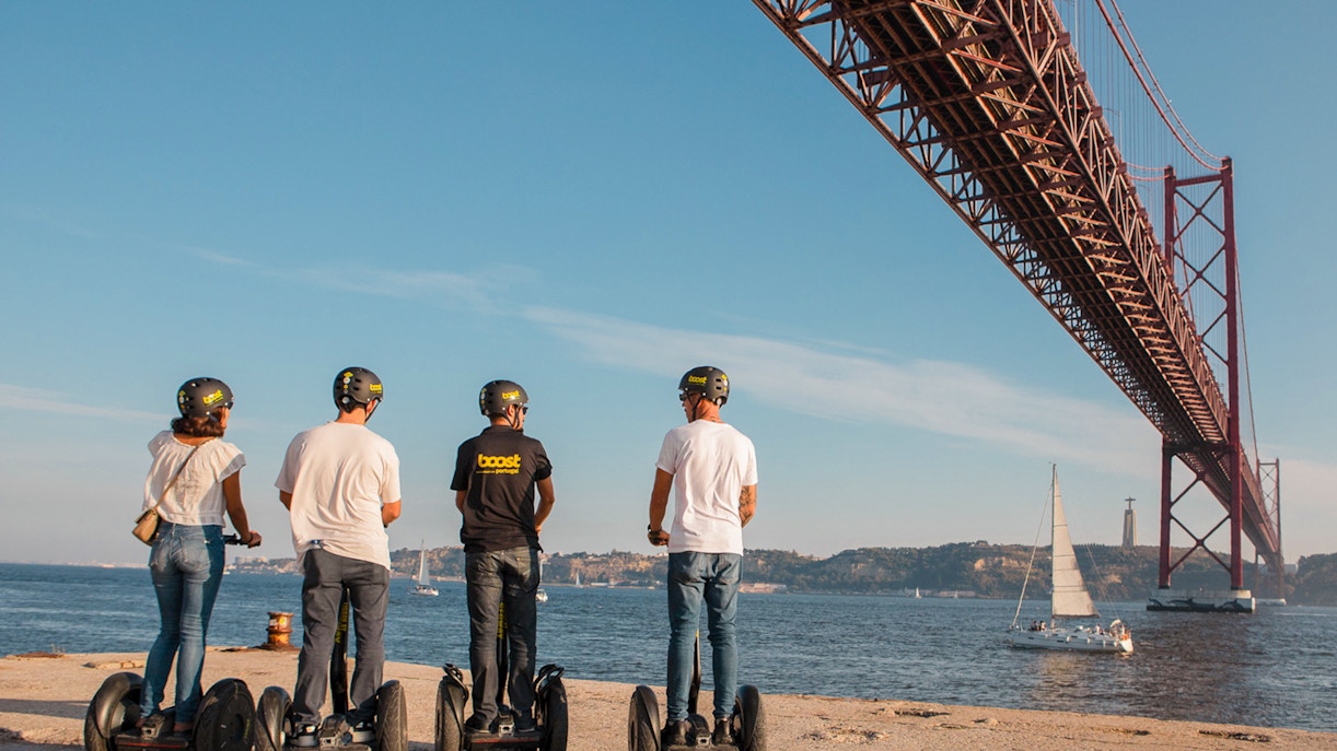 People on Segways under a large bridge in Alfama, Lisbon, with a sailboat on the river.