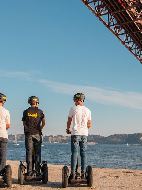 People on Segways under a large bridge in Alfama, Lisbon, with a sailboat on the river.