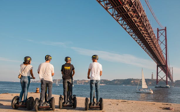 People on Segways under a large bridge in Alfama, Lisbon, with a sailboat on the river.