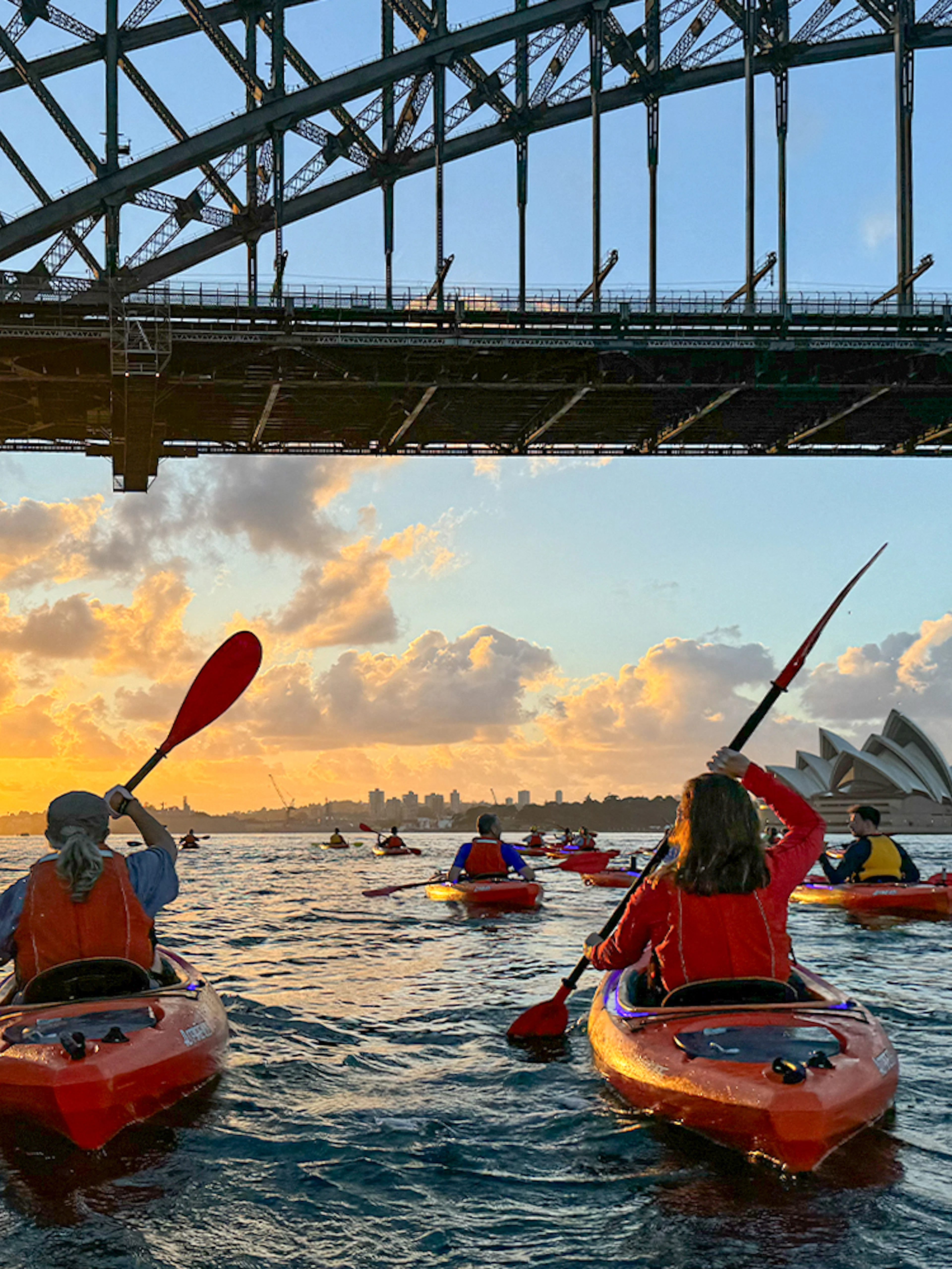 Kayakers paddling at sunrise under Sydney Harbour Bridge with Opera House view.