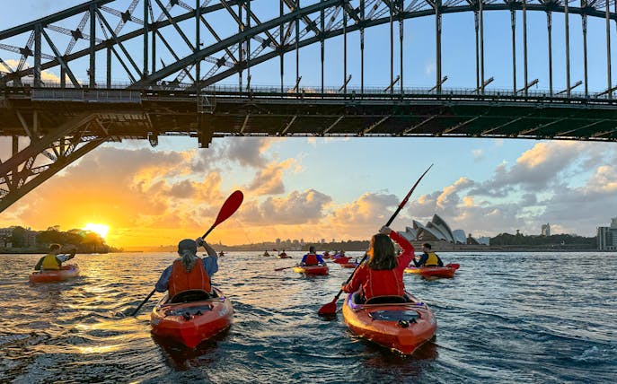 Kayakers paddling at sunrise under Sydney Harbour Bridge with Opera House view.
