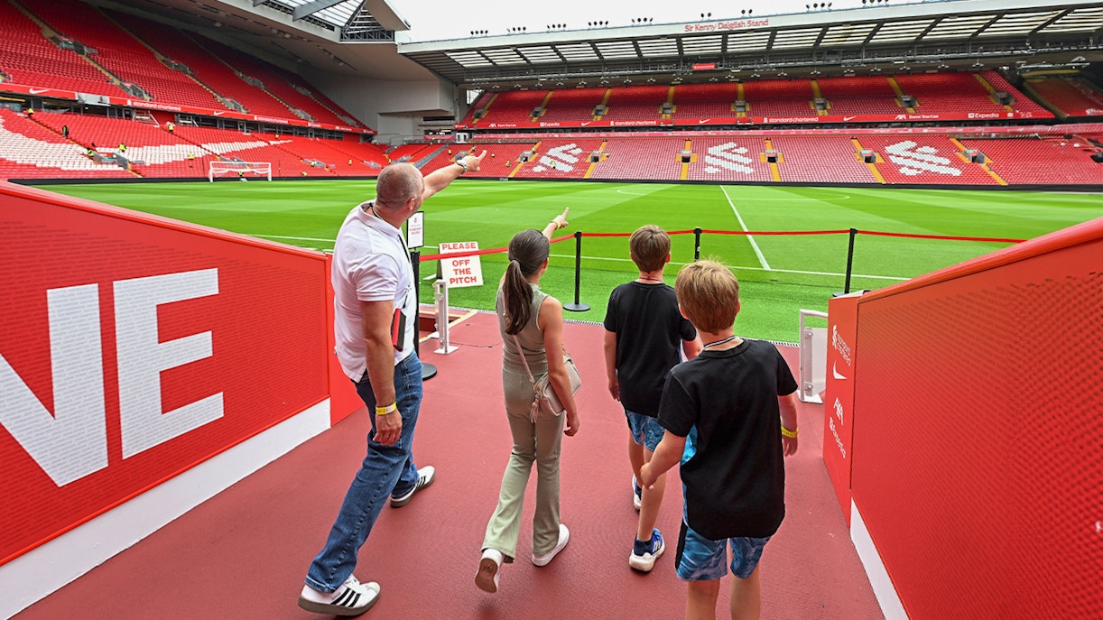 Visitors touring Anfield Stadium, home of Liverpool FC, with a guide pointing at the field.