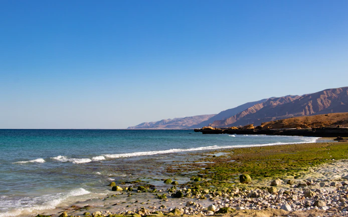 Fins Beach shoreline with rocky terrain and distant mountains near Muscat, Oman.
