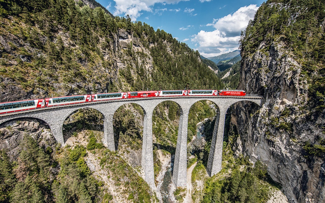Glacier Express crossing the Landwasser Viaduct in Graubünden, surrounded by forested mountains.