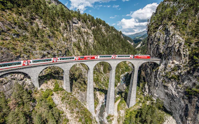 Glacier Express crossing the Landwasser Viaduct in Graubünden, surrounded by forested mountains.