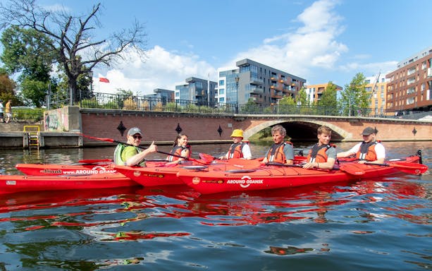 Kayakers paddling on a river in Gdansk with modern buildings and a bridge in the background.
