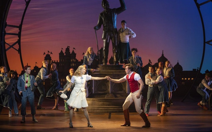 Cast performing a scene from Wicked the Musical on stage with a statue backdrop.