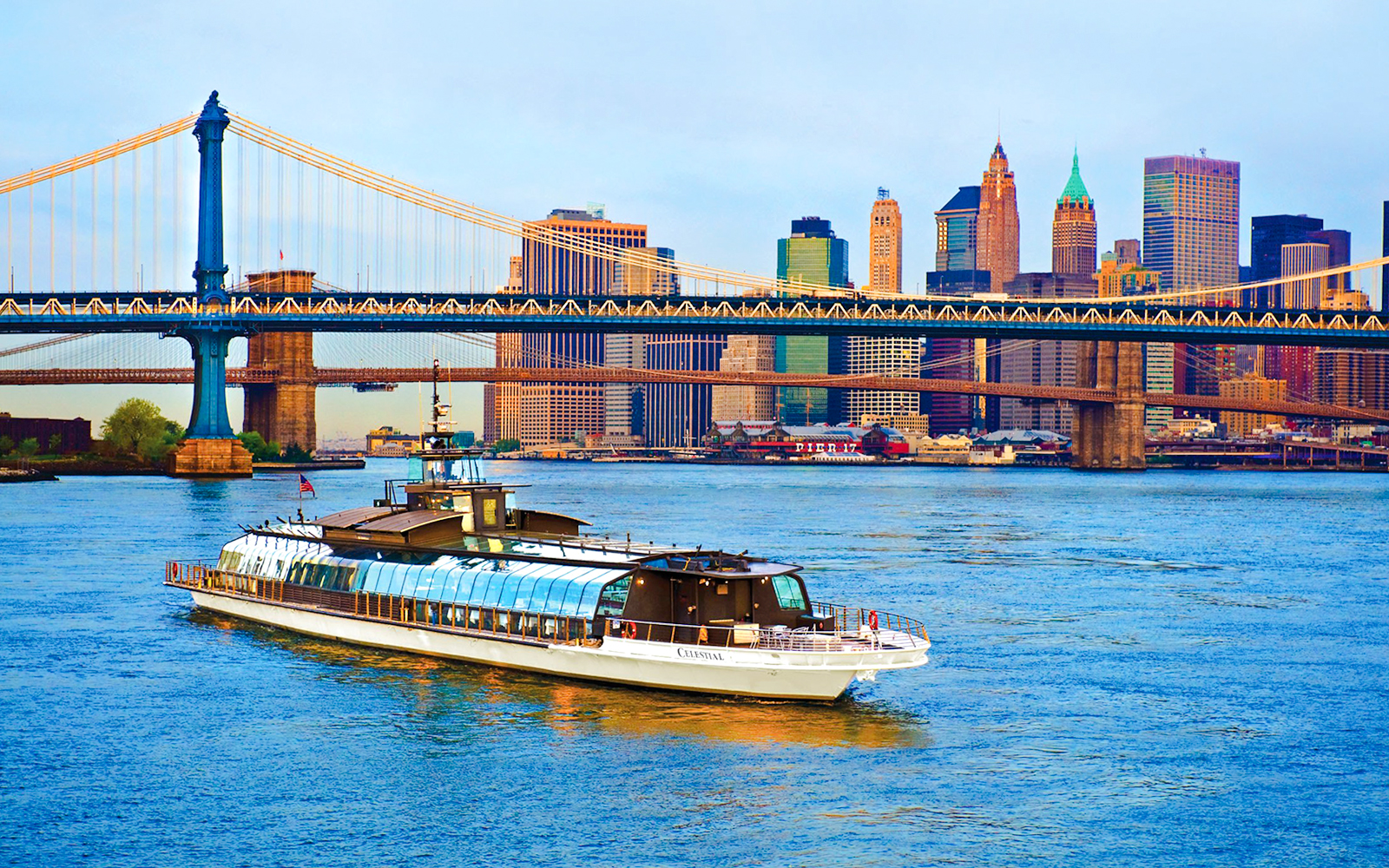 Luxury glass boat cruising under Manhattan Bridge with New York City skyline.