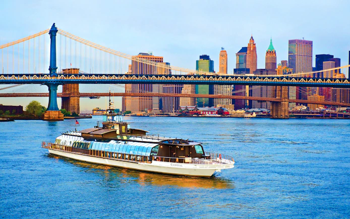 Luxury glass boat cruising under Manhattan Bridge with New York City skyline.