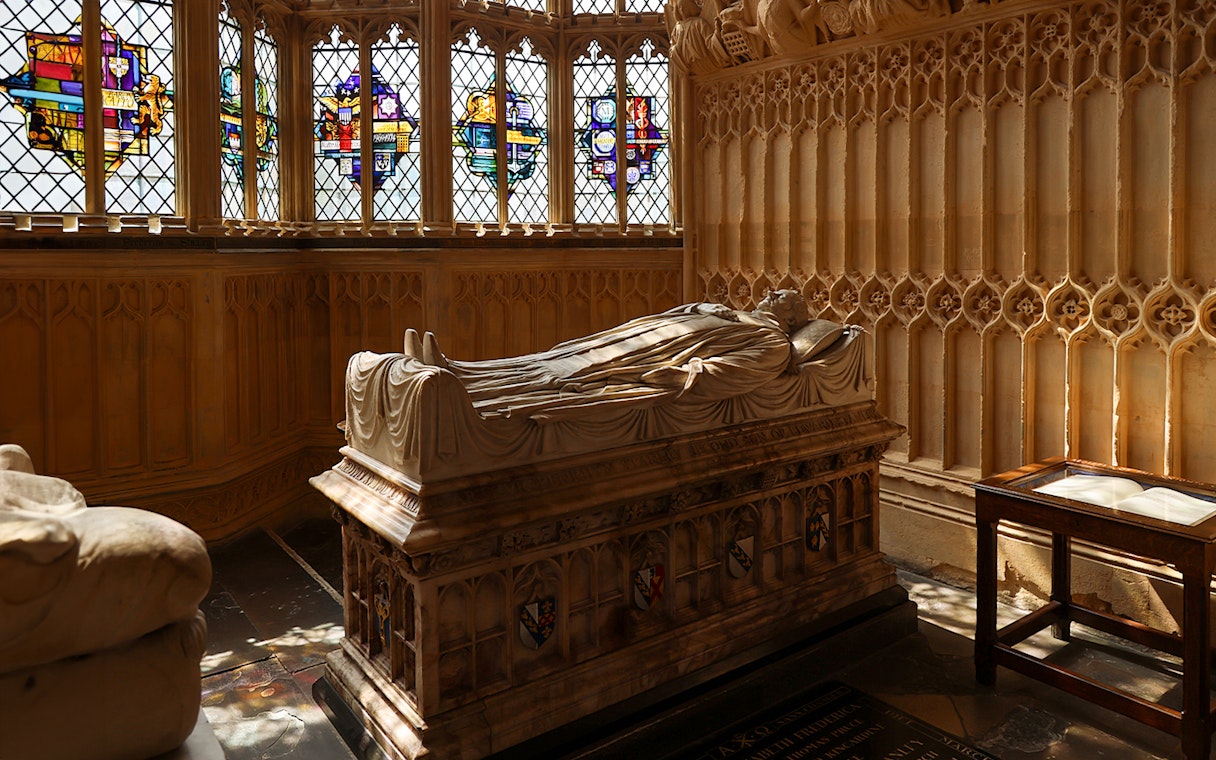 Tomb with effigy inside Westminster Abbey, London, with stained glass windows in the background.